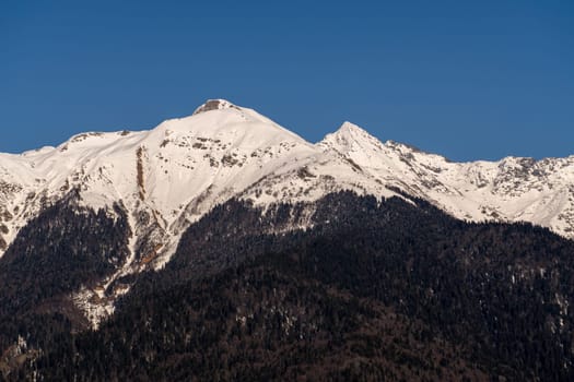 Snow-capped mountain peaks dominate a clear blue sky. Shadows cast on lower, dark forested slopes. A sunlit, winter mountain range scene.
