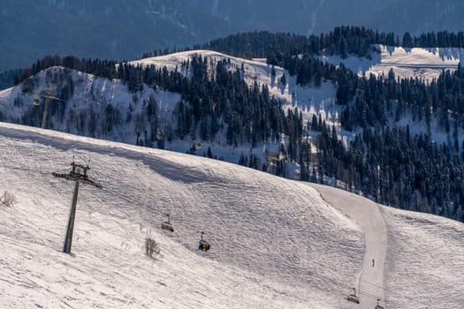 Snow-covered mountain slope with a ski lift and sparsely populated ski runs. Trees line the hillsides under a clear sky. Winter landscape.