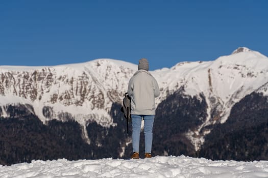 Person in light gray jacket and jeans stands on snowy mountain peak, gazing at snow-covered mountain range under a clear blue sky. Back view.