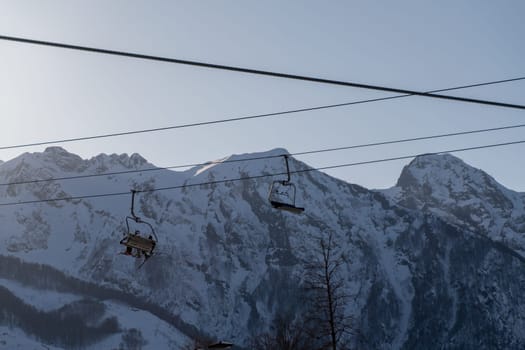 Two empty ski lift chairs hang against a backdrop of snow-covered mountains under a pale sky. Wires stretch across the scene.