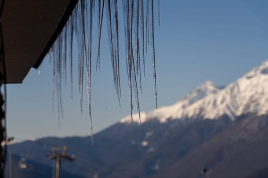 Numerous icicles hang from a roof's edge, a stark contrast to the snow-capped mountains in the background under a clear blue sky.