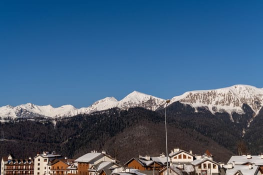 Snow-capped mountains rise majestically behind a picturesque village, their peaks sharply defined against a vibrant blue sky. Winter scene; snow covers rooftops.