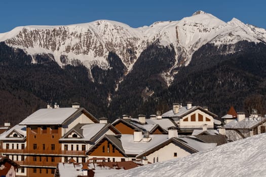 Snow-covered village nestled beneath a towering, snow-capped mountain range on a sunny day. Alpine architecture dominates the foreground.
