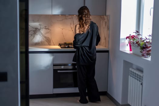 Woman in black pajamas cooks on a modern kitchen stove. Back view. Built-in oven and window visible.