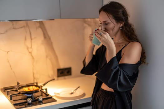Woman in silk robe stands in kitchen, sipping from a small cup. A frying pan with food sits on the stove. Morning routine in a modern kitchen.