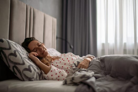 Woman sleeps soundly in bed, curled slightly, hand resting on stomach. Grey bedding, heart-patterned pillow. Sunlight streams through sheer curtains.