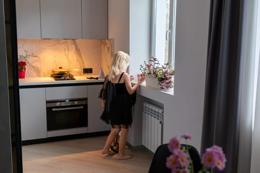 Blonde woman in black dress stands barefoot in modern kitchen, near window with flowers, looking outside. Countertop shows food prep.