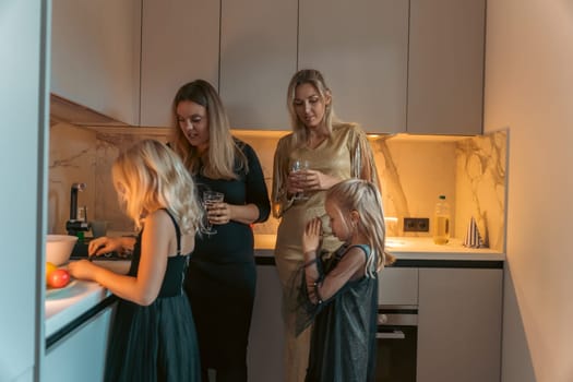 Two women and two girls in a modern kitchen. One girl cuts fruit at the counter while the others watch. Warm lighting illuminates the scene.