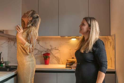 Two women in a modern kitchen one reaches for high cabinets, the other observes, possibly pregnant.