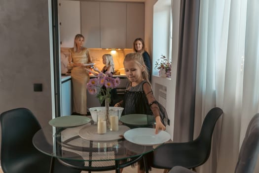 Woman sets a table in a modern dining area as two others prepare food in the adjacent kitchen. Glass table, dark chairs, and a floral arrangement are visible.