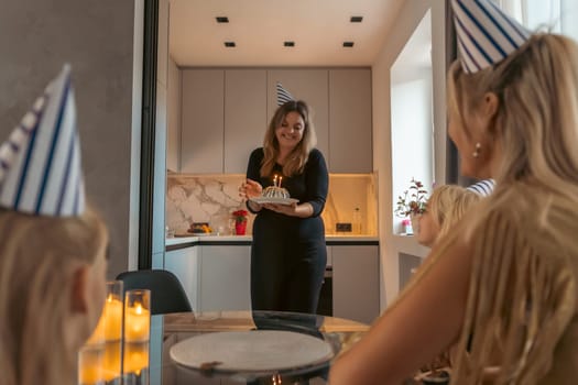 Woman presents birthday cake to two women wearing party hats in a modern apartment. Celebration in progress.