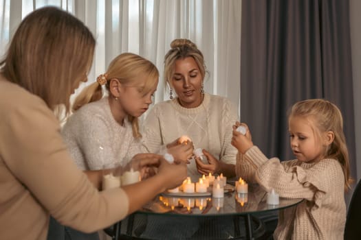Two women and two girls sit around a glass table, lighting numerous small candles. A warm, intimate family scene.