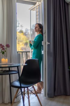 Woman in teal dress stands on balcony, holding a mug, gazing at a view. A small table and chair are nearby, inside an apartment.