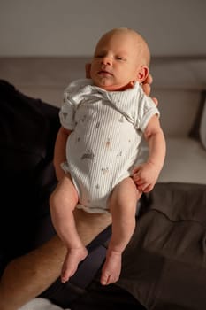 A small baby in a cozy indoor setting, gently held in a hand, wears a soft onesie and has a calm, tender expression.