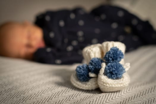 Soft white booties with blue pom poms rest on a blanket while a sleeping baby lies peacefully in the background of a serene indoor space