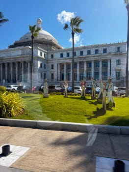 Puerto Rico, San Juan November 22. 2023 Marble Capitol under blue sky, with palm trees and vibrant cutout figures displayed on the green lawn.
