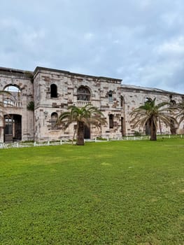 Weathered stone ruins of a historic colonial-era building in Bermuda, surrounded by green lawn and palm trees under cloudy sky.