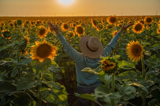 Woman in hat stands in a sunflower field at sunset, arms raised, enjoying the golden light. Bright yellow flowers surround her.