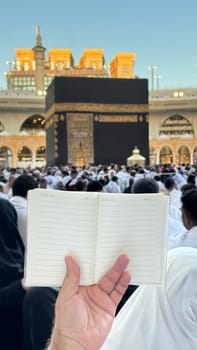 A pilgrim holds a small notebook with a heart doodle in front of the Kaaba in Mecca, Saudi Arabia. The sacred site is surrounded by worshippers performing Hajj or Umrah under a clear blue sky.