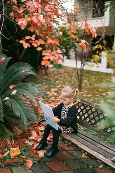 Little girl looks at the cover of a book while sitting on a bench in an autumn park. High quality photo