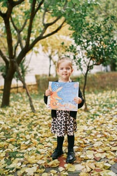 Little girl with a colorful book in her hands stands in the autumn garden. High quality photo