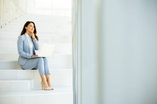 Pretty young business woman sitting on the stairs on office hallway and working on laptop
