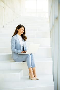 Pretty young business woman sitting on the stairs on office hallway and working on laptop