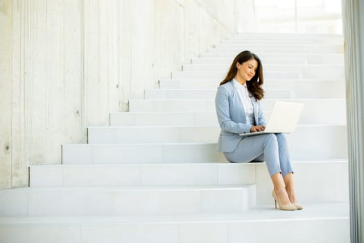 Pretty young business woman sitting on the stairs on office hallway and working on laptop