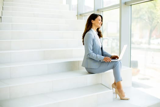 Pretty young business woman sitting on the stairs on office hallway and working on laptop