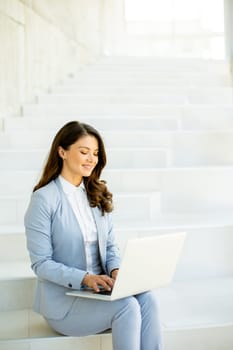 Pretty young business woman sitting on the stairs on office hallway and working on laptop