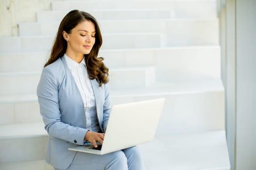 Pretty young business woman sitting on the stairs on office hallway and working on laptop