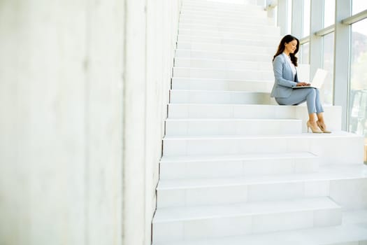 Pretty young business woman sitting on the stairs on office hallway and working on laptop