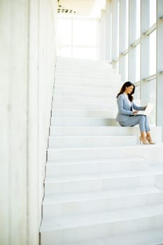 Pretty young business woman sitting on the stairs on office hallway and working on laptop