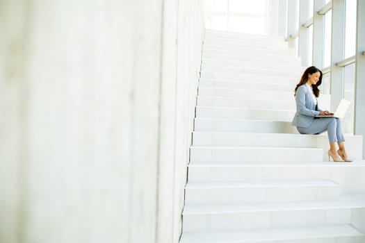 Pretty young business woman sitting on the stairs on office hallway and working on laptop
