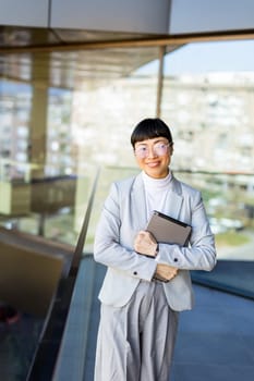 Standing in a stylish office space, a Japanese professional woman holds a tablet close while smiling. Natural light floods through large windows, highlighting their confident presence.