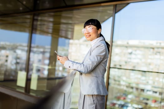 Bright sunlight illuminates a confident Asian woman in a chic suit, smiling as she engages with her smartphone against a backdrop of contemporary architecture in a bustling urban environment.