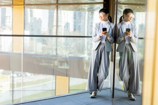 A joyful professional Asian woman dressed in a stylish gray suit smiles widely while holding her phone. She stands in a modern office with large windows reflecting a vibrant cityscape outside.