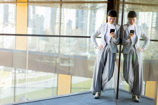 A focused professional Japanese woman dressed in a stylish business suit scrolls through her smartphone. She stands near a large glass window reflecting a vibrant urban landscape outside.