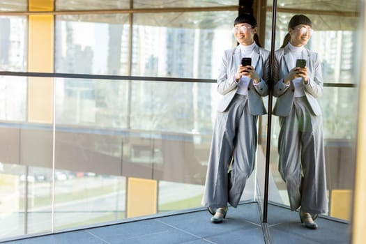 A joyful professional Asian woman dressed in a stylish gray suit smiles widely while holding her phone. She stands in a modern office with large windows reflecting a vibrant cityscape outside.