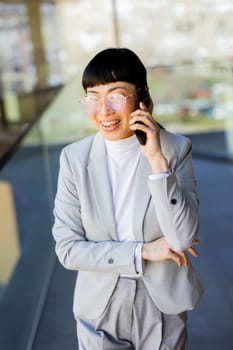 A professional Asian woman in a stylish suit chats joyfully on her phone within a sleek office building with large windows. Her bright smile reflects her positive energy and success.
