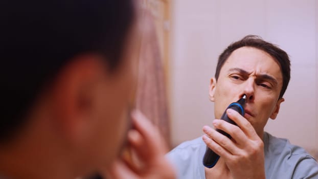 Caucasian man using a small electric trimmer to remove nose hair while looking in the mirror in his bathroom