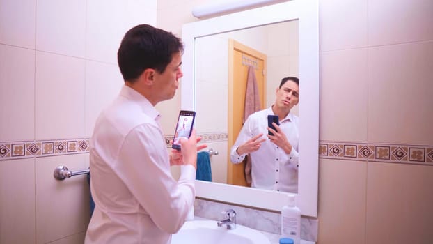 Narcissistic businessman taking pictures of himself with smartphone in bathroom mirror, adjusting hair and clothes