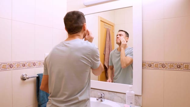 Man performs a detailed facial cleansing routine, using cotton pads and facial products in a tiled bathroom, promoting skincare habits