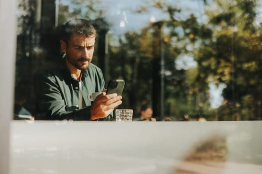 In the warm sunlight of a vibrant cafe, a man sits at a table, engrossed in his smartphone, reflecting on modern life amidst the lively atmosphere.