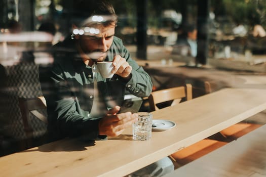 Sitting at a wooden table, a man enjoys his coffee while using mobile phone and gazing out a window, capturing the essence of solitude and reflection in a warm cafe atmosphere