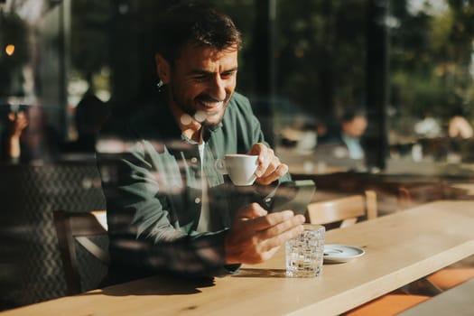 Sitting at a wooden table, a man enjoys his coffee while using mobile phone and gazing out a window, capturing the essence of solitude and reflection in a warm cafe atmosphere