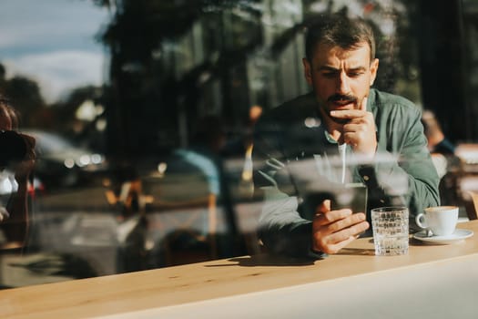 Sitting at a wooden table, a man enjoys his coffee while using mobile phone and gazing out a window, capturing the essence of solitude and reflection in a warm cafe atmosphere