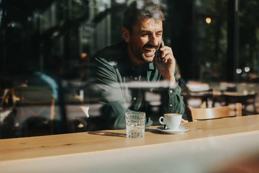 Sitting at a wooden table, a man enjoys his coffee while using mobile phone and gazing out a window, capturing the essence of solitude and reflection in a warm cafe atmosphere
