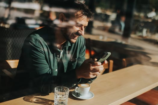 Sitting at a wooden table, a man enjoys his coffee while using mobile phone and gazing out a window, capturing the essence of solitude and reflection in a warm cafe atmosphere