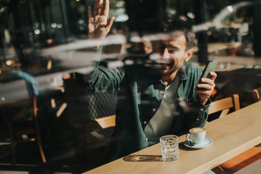 Sitting at a wooden table, a man enjoys his coffee while using mobile phone and gazing out a window, capturing the essence of solitude and reflection in a warm cafe atmosphere